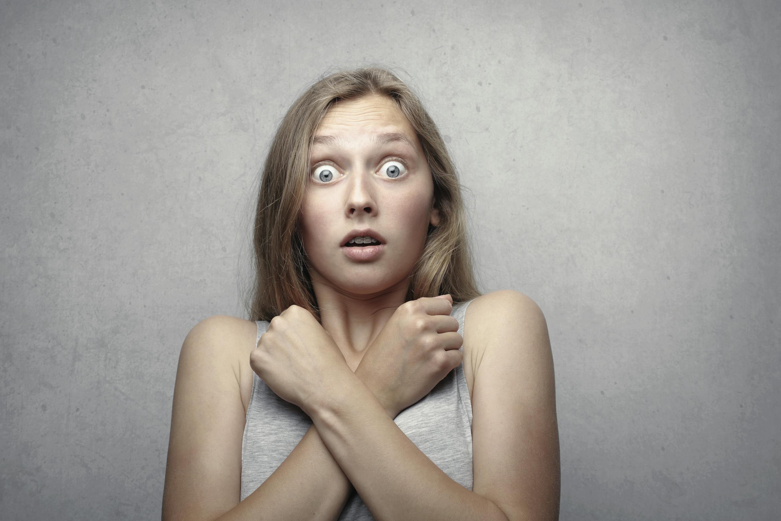 Portrait of a surprised woman with wide eyes and crossed arms against a gray background.